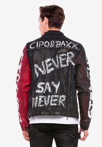 Zwarte denimjas met rode mouwen, voorzien van witte tekst en grafische ontwerpen. Zilveren studs op de schouders. Ritsluiting aan de onderkant.