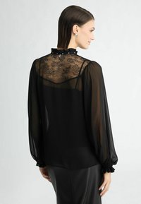 Mustvalge blouse, millel on läbipaistev pits seljaosa, volangiga kaelus ja puffvarrukad, millel on kogutud mansetid. Sile kangas annab kerge tekstuuri.