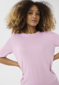 Gaiši rozā, īsām piedurknēm adīts tops ar apaļu kakla izgriezumu. Gluda tekstūra un brīvs piegriezums, ar sānu šķēlumiem papildu detaļām.