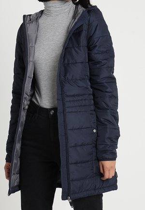 Vrouw draagt een marineblauw doorgestikt jasje over een grijze coltrui en zwarte broek tegen een effen achtergrond.