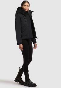 Black puffer jacket with a high collar and hood, featuring front zippers and side pockets, paired with black leggings and chunky black boots.