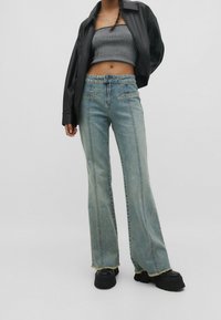 Jeans acampanados de un azul claro con un dobladillo deshilachado, que cuentan con bolsillos frontales, detalles de costura sutiles y un corte relajado. Combinados con una chaqueta de cuero negra.