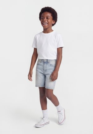 Mosolygó gyermek afro hajjal, fehér pólót, világos farmer rövidnadrágot, fehér zoknit és fehér tornacipőt visel, és egy egyszínű háttér előtt áll.