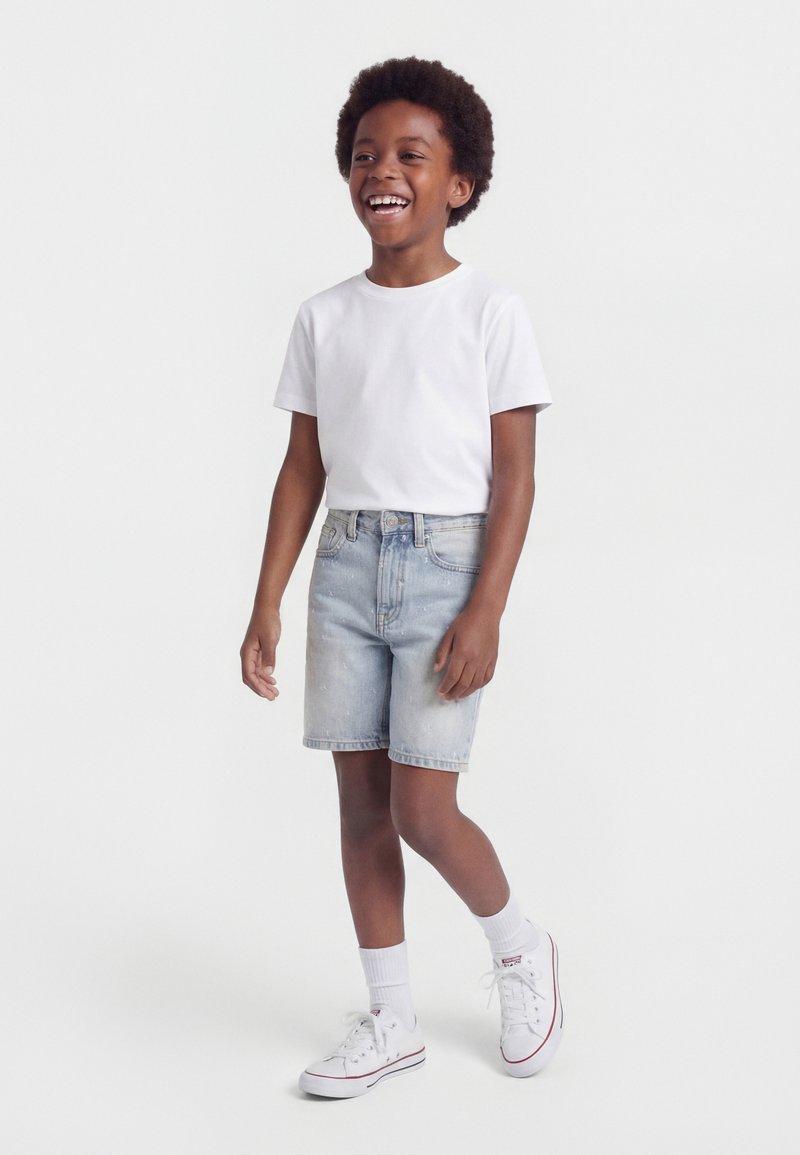 Χαμογελαστό παιδί με αφρομαλλιά, που φοράει λευκό μπλουζάκι, ανοιχτόχρωμο τζιν σορτς, λευκές κάλτσες και λευκά αθλητικά παπούτσια, στέκεται μπροστά σε απλό φόντο.