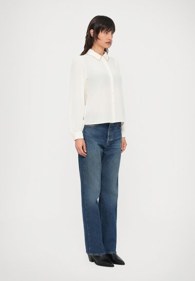 Λευκό μπλουζάκι μακρυμάνικο με κουμπιά μπροστά, γιακά με αιχμή, συνδυασμένο με ψηλόμεσα μπλε τζιν και μαύρα μποτάκια με τακούνι.