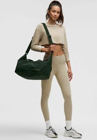 Oliwkowa torba podróżna z teksturowaną powierzchnią, kieszenią na zamek błyskawiczny i regulowanym paskiem, w połączeniu z beżowym topem w wersji cropped i legginsami.
