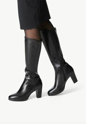 Czarne skórzane kozaki za kolano na słupku, z bocznymi zamkami, noszone z czarnymi pończochami i krótkimi spodniami na białym tle.