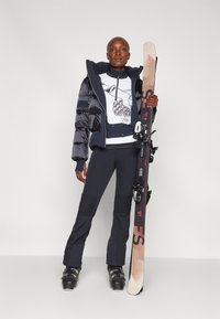 Skioutfit met een dikke zwarte jas, wit graphic longsleeve shirt, zwarte skibroek met textuurpanelen en ski's met bindingen.