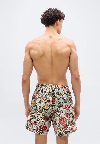 Νεαρός άνδρας με σγουρά μαλλιά, γυμνή πλάτη, φορώντας πολύχρωμο σορτς με ανθισμένο σχέδιο, στέκεται μπροστά σε έναν απλό φωτεινό φόντο.