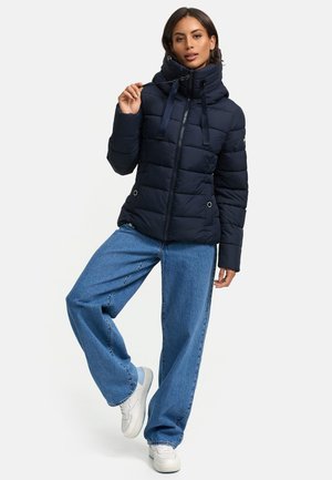 Veste matelassée navy avec un col haut et des cordons de serrage, dotée d'un design matelassé et d'une fermeture éclair, associée à un jean bleu à jambe large et des baskets.