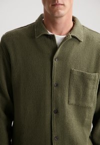 Cămașă texturată verde, cu guler, având un buzunar aplicat și nasturi negri. Fabricată din material țesut.