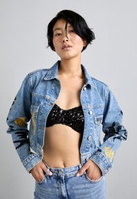 Asmuo su trumpais juodais plaukais, vilkintis mėlyną džinsinį švarką su juodais ir auksiniais raštais, juodu blizgučiuotu liemenėliu ir mėlynomis džinsomis, rankas kišenėse.