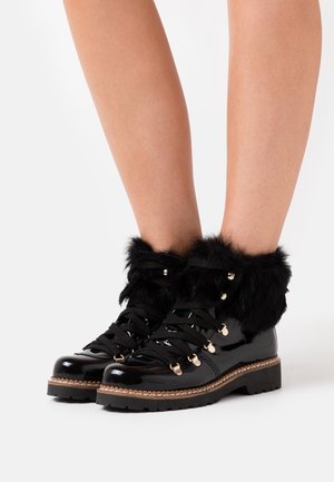 Bottes en cuir verni noir avec des poignets en fausse fourrure, dotées de lacets noirs et d'accents en métal doré. Des semelles épaisses et texturées complètent le design.