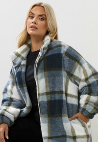 Fleece oversized jacka i blått, vitt och grönt rutmönster med hög krage, dragkedja framtill och sidofickor för ökad funktionalitet.