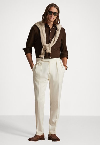 Καφέ πουκάμισο με κουμπιά, μπεζ πουλόβερ γύρω από τον λαιμό, λευκά πλισέ παντελόνια και καφέ δερμάτινα παπούτσια. Αξεσουάρ συμπεριλαμβάνουν γυαλιά ηλίου.