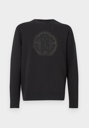 Svart genser laget av mykt stoff, med en teksturert sirkulær logo i midten omgitt av dekorative detaljer. Lange ermer.