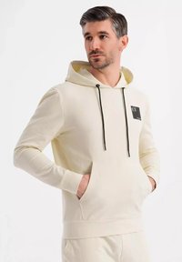 Cremefarbener Hoodie aus weichem Stoff, mit einer Fronttasche, einer Kordelzugkapuze mit schwarzen Akzenten und einem kleinen Logo-Patch auf der Brust.