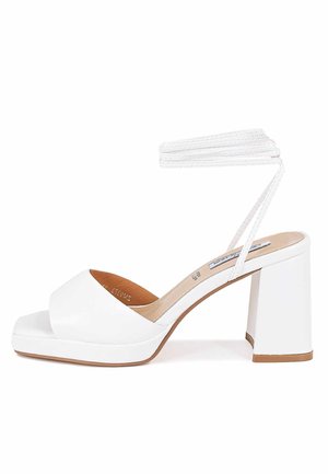 Hvide højhælede sandaler med en glat læderfinish, åben tå, bred rem over fronten og en blokhæl. Har en flettet ankelrem.