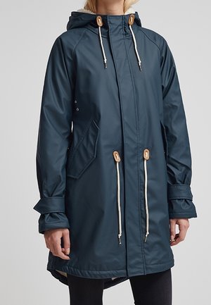 Imperméable à capuche bleu marine avec cordons blancs et boutons en bois, poches à rabat et sangles de poignets ajustables porté par une personne.