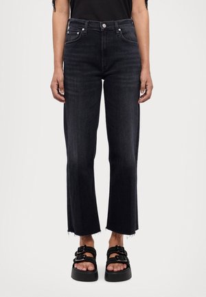 Personne portant un jean noir taille haute à jambes droites avec des ourlets bruts et des sandales à plateforme noires à doubles boucles.