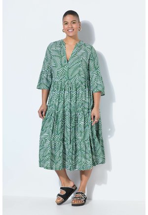 Vrouw die een midi-jurk draagt met een groen-wit bladpatroon, driekwart mouwen en zwarte slip-on sandalen met witte bies, glimlachend.