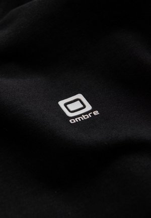 Tessuto nero con un logo bianco ben visibile, contornato da un quadrato e con la parola "ombre" in lettere minuscole, dalla texture liscia.