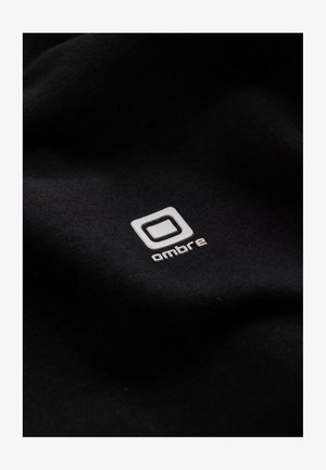 Czarna tkanina z wyraźnym białym logo w kwadratowej ramce oraz napisem "ombre" napisanym małymi literami, gładka tekstura.