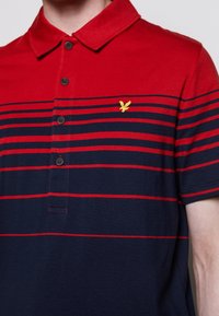 Polo shirt a righe rosse e blu con colletto a bottoni e un piccolo logo ricamato giallo sul lato sinistro del petto. Tessuto di cotone, maniche corte.