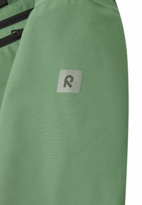 Grønt soft-shell stof med en mat tekstur, prydet med et sølvfarvet firkantet logo. Indeholder sorte lynlåslukninger til lommer.
