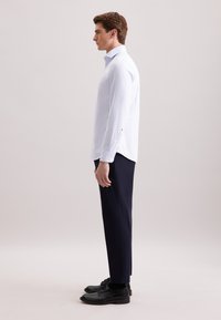 Camicia bianca a maniche lunghe con colletto a bottoni, abbinata a pantaloni blu scuro e scarpe nere. La camicia presenta un orlo dritto e dettagli con bottoni.