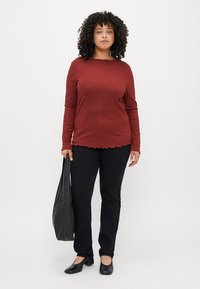 Ribbed, dlhý rukáv burgundský top s vykrojeným lemom, skombinovaný s čiernymi nohavicami a plochými topánkami. Modelka drží čiernu tašku.