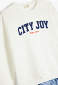 Hvid sweatshirt med "CITY JOY" i marineblå bogstaver og "Happy Moves" i orange. Har rund hals og ribbede manchetter. Blød stofstruktur.