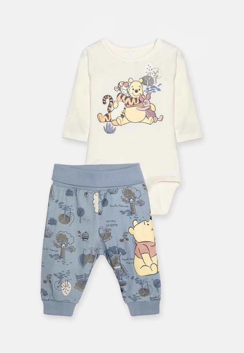 Bodysuit blanc à manches longues avec des personnages colorés de Winnie l'Ourson. Pantalon bleu avec des motifs de forêt ludiques et une taille élastique.