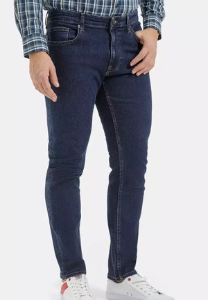 Mężczyzna noszący ciemnoniebieskie dopasowane jeansy, białe trampki i zieloną koszulę w kratę, stojący na białym tle.