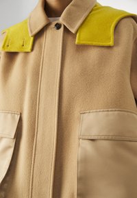 Manteau en laine beige avec large col, accents aux épaules jaune vif, bouton frontal et grandes poches à rabat beiges.