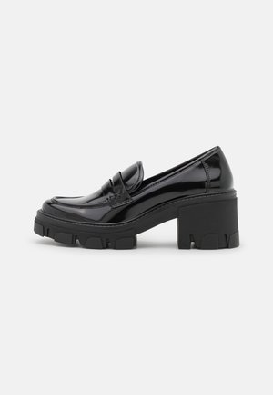 Loafer à talon épais en cuir verni noir avec une semelle crantée épaisse et une sangle décorative à l'avant, présenté en profil latéral sur un fond uni.