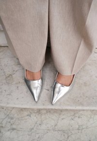 Zilveren puntige schoenen met een gladde metallic afwerking, geaccentueerd door een open vierkante uitsnijding aan de bovenkant, gecombineerd met lichtbeige, gestructureerde broeken.