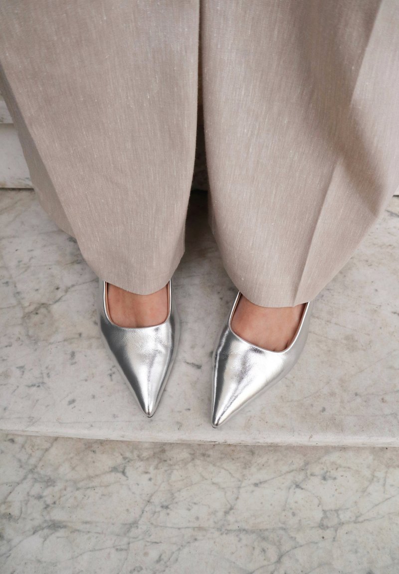 Zilveren puntige schoenen met een gladde metallic afwerking, geaccentueerd door een open vierkante uitsnijding aan de bovenkant, gecombineerd met lichtbeige, gestructureerde broeken.