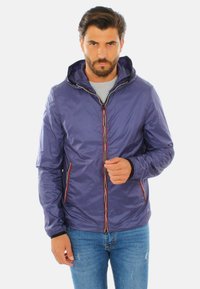 Giacca leggera viola con cappuccio, dotata di una zip frontale, dettagli rossi e bianchi e due tasche laterali. Indossata sopra una maglietta grigia e jeans.