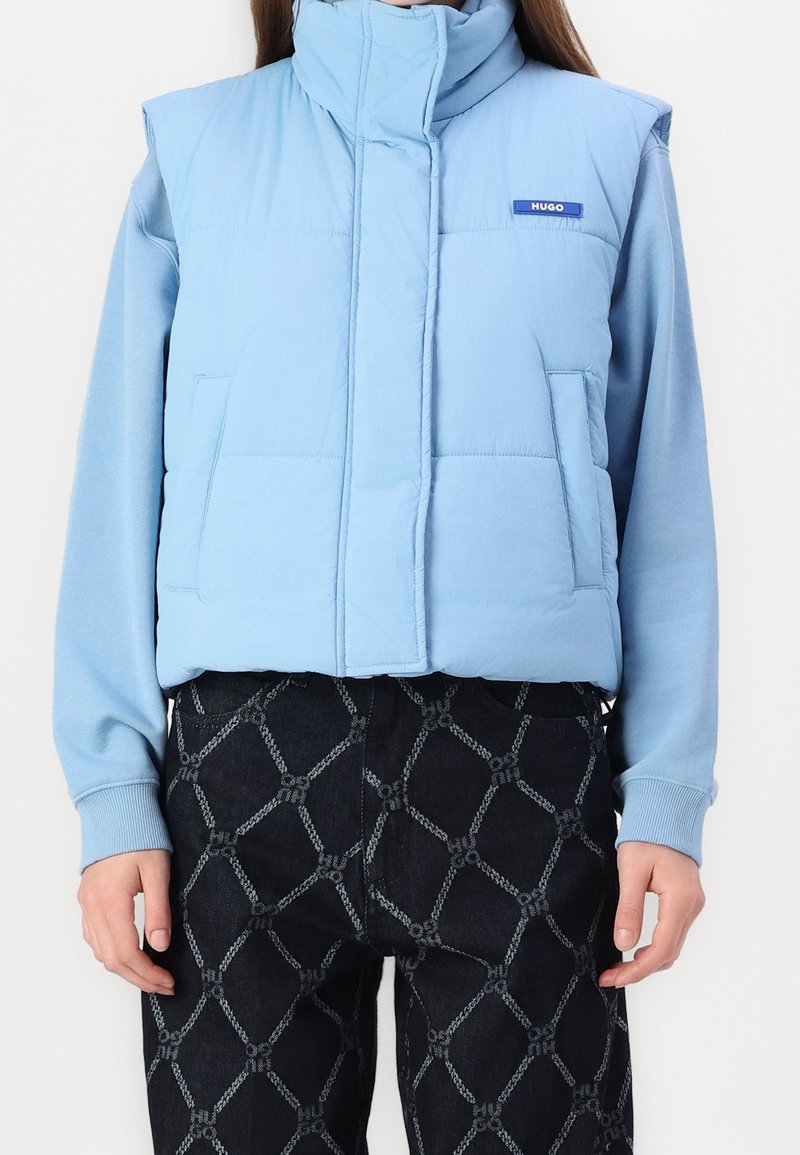 Helles blaues Pufferwesten mit hohem Kragen, zwei Fronttaschen und Logo-Detail. Getragen über einem hellblauen Langarmshirt und gemusterten dunklen Jeans.