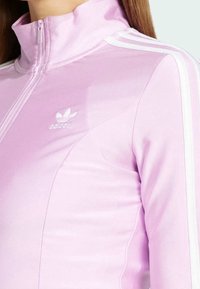 Närbild av en person som bär en ljusrosa Adidas zip-up jacka med vita ränder på axeln och Adidas-logotypen på bröstet.