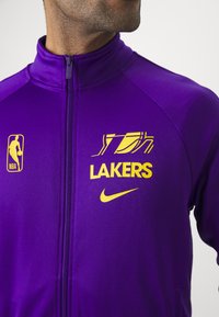 Fato de treino roxo da Nike com o logótipo amarelo "Lakers" e emblema da NBA, fecho de zíper e textura de tecido suave.