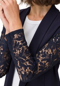 Blazer azul marino con largas mangas de encaje que presentan un intrincado patrón floral. El interior se combina con una blusa blanca de canalé.