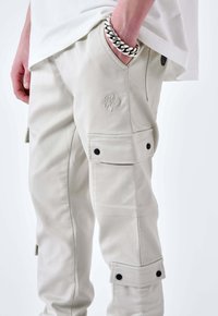 Persona che indossa pantaloni cargo beige con tasche a patta e un bracciale d'argento, mano in tasca contro uno sfondo bianco.