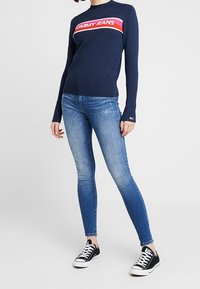 Kvinna iklädd en marinblå Tommy Jeans-tröja, ljusblå skinny jeans och svarta låga sneakers står mot en vit bakgrund.