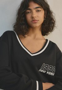 Černá mikina s výstřihem do V a bílými pruhovanými detaily. Obsahuje vyšívaný text "1990 NEW YORK." Měkká tkanina s uvolněným střihem.