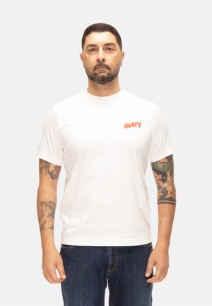 Hombre con cabello corto y oscuro y barba, vistiendo camiseta blanca Autry y vaqueros azules, mostrando tatuajes coloridos en ambos antebrazos, de pie contra fondo blanco.