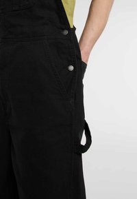 Salopette en denim noir avec poches latérales, accents de boutons en métal argenté, une boucle utilitaire et une coupe décontractée. Tissu texturé et durable.