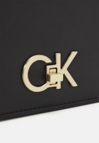 Schwarze, strukturierte Lederhandtasche mit einem auffälligen goldenen "CK"-Logo und einem rechteckigen Verschlussmechanismus am Logo.