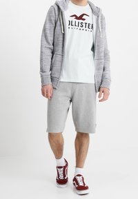 Mężczyzna w szarej bluzie z zamkiem, białej koszulce Hollister, szortach w kolorze szarym, białych skarpetkach oraz czerwonych trampkach idzie na tle białego tła.